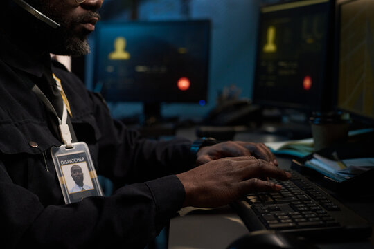 Black man working as dispatcher typing on computer keyboard in control room, wearing headset and identification badge, with multiple monitors displaying information in background
