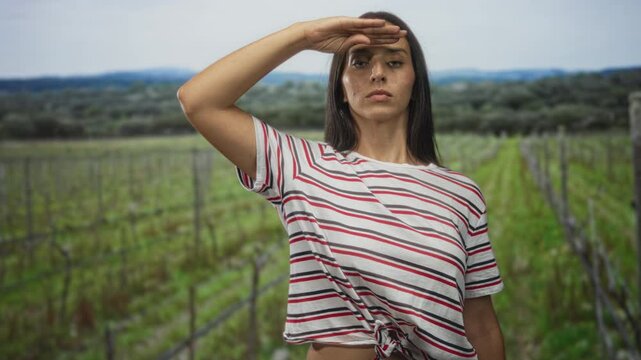 Woman hand to forehead shading eyes in forest vineyard rows with striped shirt and knotted hem exposing midriff; determination.