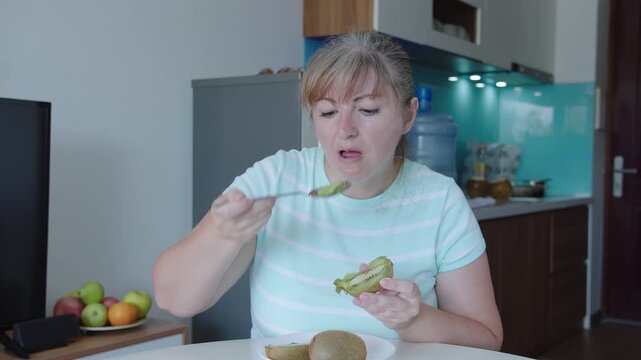 A woman eats kiwi pulp with a spoon, sits at a table, kitchen background