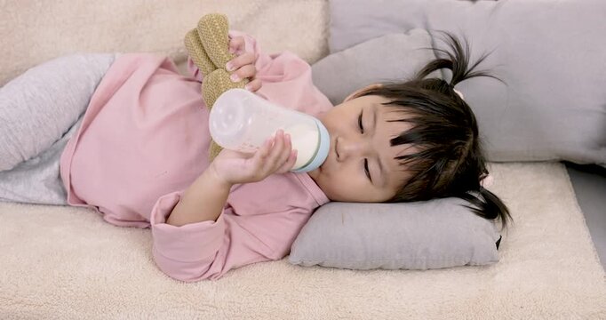 Asian preschool daughter smiling gently while relaxing on cushion holding milk bottle and plush toy showing emotional safety, early childhood independence and peaceful behavior during rest in home