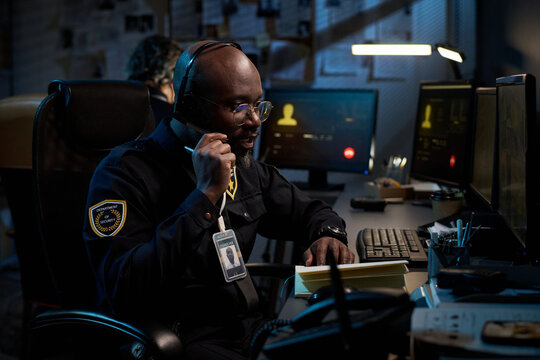 Young Black man wearing headset sitting at desk talking on phone while monitoring computer screens in dark office, visible security badge hanging from neck, working night shift