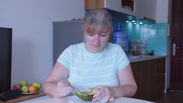 Woman eats avocado pulp with a spoon, sits at a table