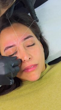 Professional artist performing eyebrow threading for a beautiful woman, creating symmetrical brows.