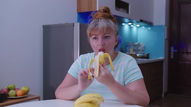 Woman peels and eats a banana sitting at a table , kitchen background