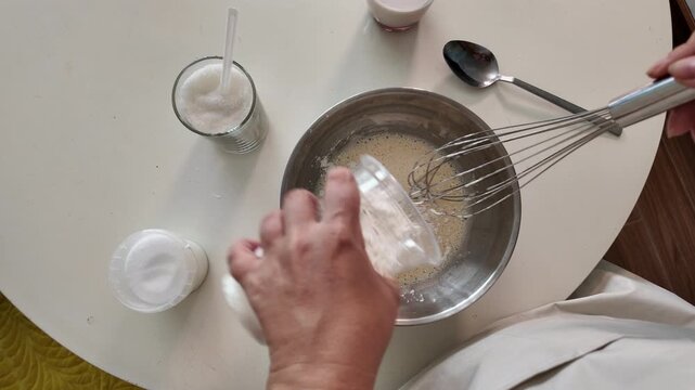 A woman pours flour into an egg mixture and whips the mixture with a whisk