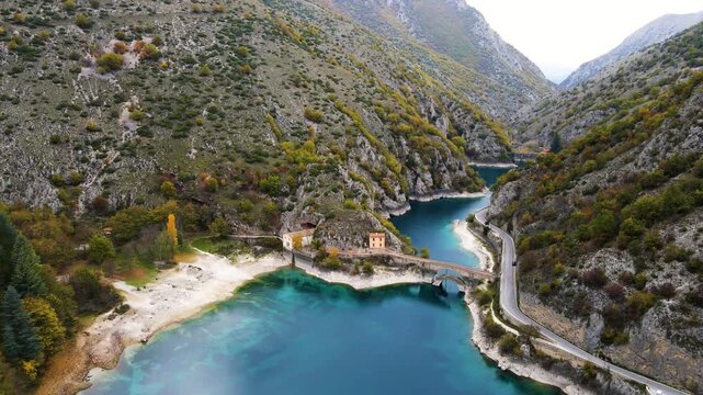 Expansive aerial view of Sagittario River gorge with historic stone bridge, vibrant turquoise water, autumn forest, and winding mountain road through dramatic limestone canyon.