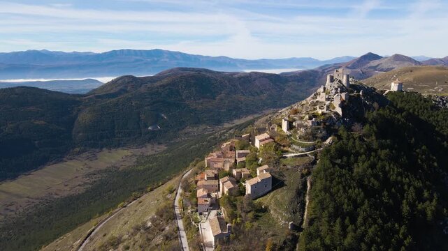 Stunning aerial of Rocca Calascio medieval hilltop village cascading down rocky ridge with fortress ruins above, valley fog below and dramatic Gran Sasso mountain range beyond.