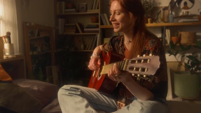Cinematic medium shot of creative young woman singing and playing guitar while sitting on bed at home writing music in sunlight, copy space