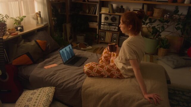 Wide angle side view shot of girl watching videos or movies online with police troops on screen while sitting on bed at home and drinking tea in morning, copy space