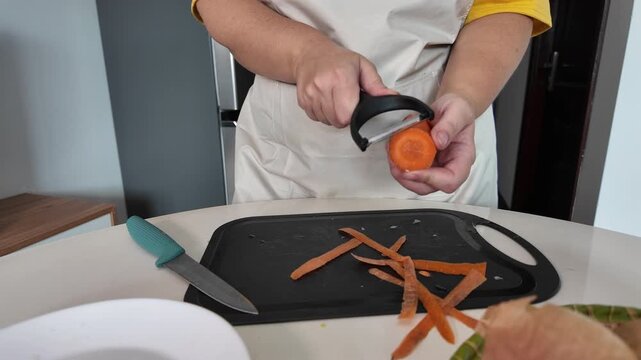 Hands of a woman who, in an apron, peels carrots with a vegetable peeler