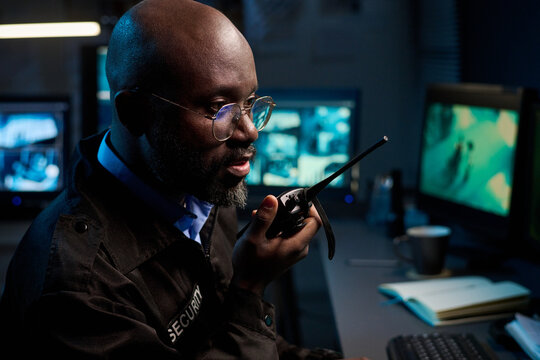 Black man wearing glasses operating walkie talkie while monitoring surveillance footage on multiple screens in security control room during night shift