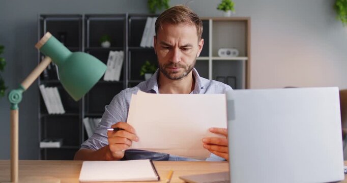 Man at laptop analyzes paperwork and report. In a tidy office, he checks documents for audit and performs thorough analysis to validate finance data and figures. Clear concept of audit analysis.