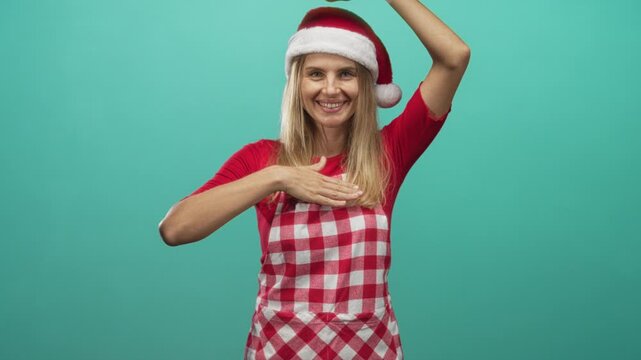 Blonde woman wearing santa hat and red gingham apron smiles with right hand on chest and left arm raised against teal studio backdrop; festive holiday cheer.