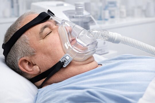 Person lying in bed wearing a CPAP mask connected to a hose, illustrating sleep apnea therapy and respiratory support in a clinical setting. Ideal for medical and healthcare awareness visuals.