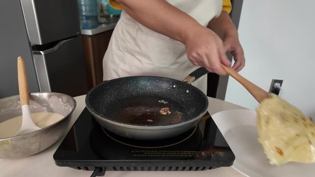 A woman in an apron frying pancakes. Close-up. Flips a pancake with a wooden spatula on an induction hob