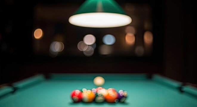 Billiard table with colorful balls under a bright green lamp in a dimly lit room.