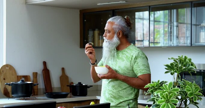 Indian mid age man eating poha while standing in home kitchen in the morning, stylish bearded male holding bowl with healthy breakfast and enjoying traditional Indian meal with spoon
