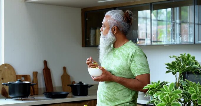 Indian mid age man eating poha while standing in home kitchen in the morning, stylish bearded male holding bowl with healthy breakfast and enjoying traditional Indian meal with spoon