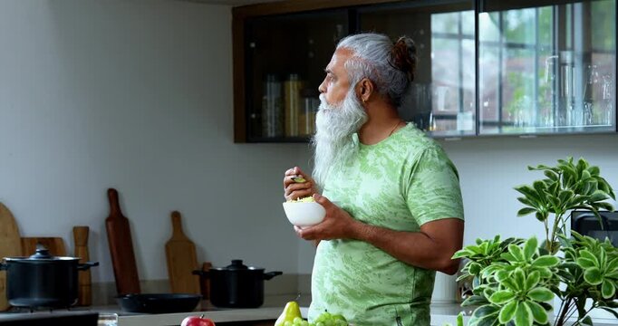 Indian mid age man eating poha while standing in home kitchen in the morning, stylish bearded male holding bowl with healthy breakfast and enjoying traditional Indian meal with spoon