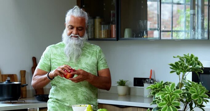 Indian mid age man holding and showing fresh red apple to camera, stylish bearded male promoting healthy eating while flexing bicep muscle and pointing at fruit standing in modern kitchen interior