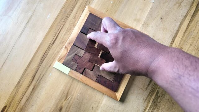 Top-down view of a person's hands solving a complex wooden brain teaser puzzle on a table. The final pieces are put into place