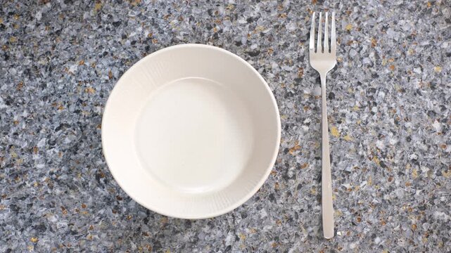 Female hand holding a fork next to an empty plate on gray background close-up top view.