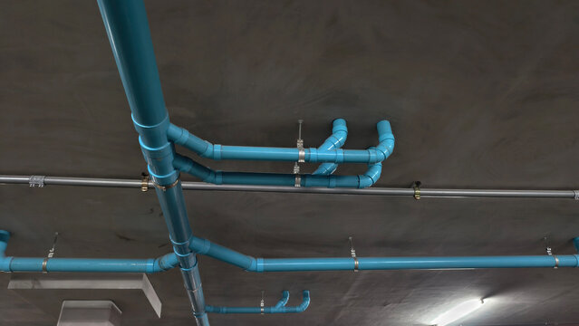 Industrial blue PVC pipes and gray conduits installed along dark concrete ceiling. A professional technical shot ideal for plumbing construction and building maintenance services
