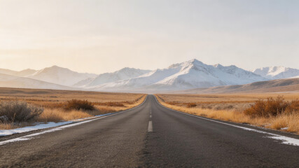 An empty asphalt highway stretches toward snow-capped mountains under a soft sunset sky, with dry grass and patches of snow lining the roadside in a vast, open valley landscape.