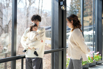 Fototapeta premium A man and a woman are holding a baby in a room with a view of a garden. Scene is warm and loving, as the family is spending quality time together