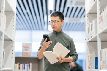 Fototapeta premium Young man in a library holding an open book while checking his smartphone