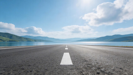 A long, empty highway stretches toward a blue lake and distant mountains under a bright sky with scattered clouds. Sunlight enhances the serene, natural landscape ideal for travel and adventure.
