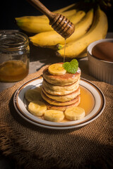 Stack of fluffy homemade banana pancakes topped with fresh slices, mint leaf, and golden honey dripping from a wooden dipper. Rustic breakfast concept on burlap background with natural sunlight. © Chayut