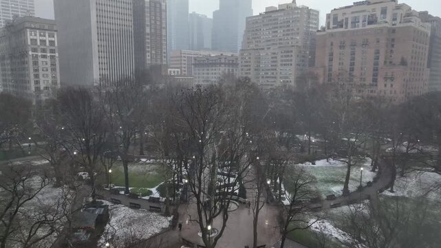 Approaching aerial shot over Rittenhouse Square in Philadelphia, USA during light snowfall. Downtown skyscrapers rise through gray winter haze as drone moves forward above park&rsquo;s bare trees.