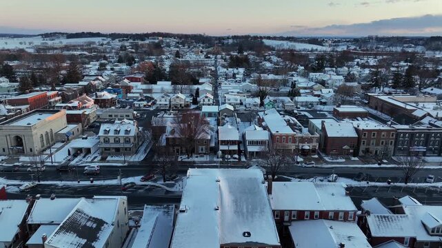Aerial sunset view over historic Manheim, Pennsylvania, covered in fresh snow. Colorful brick buildings and classic American houses line streets, with white rooftops glowing in winter light. Flyover.