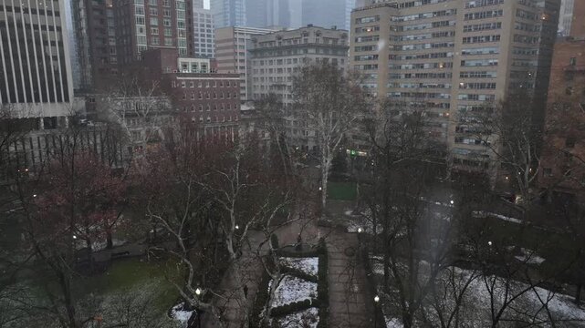 Aerial view of light snow flurries drifting over Rittenhouse Square in Philadelphia, Pennsylvania. Winter trees frame park as surrounding high-rise buildings fade into a soft, snowy haze. Descend.