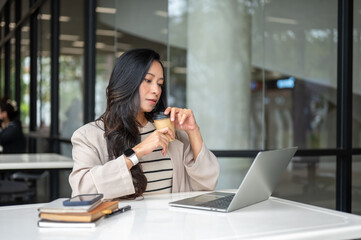 Fototapeta premium Asian woman holding coffee looking at laptop aside book and phone sitting at table in cafe building.