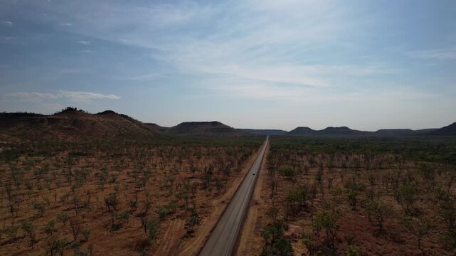 Drone captures a van driving along empty Victoria Highway in Northern Territory, surrounded by vast bushland, red dirt and distant mountain ranges.