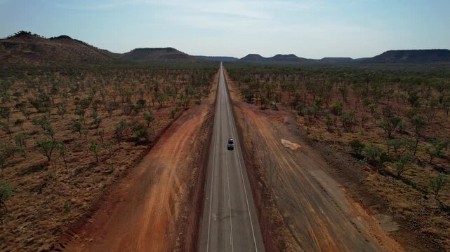 Van drives along empty Victoria Highway in Northern Territory, surrounded by vast bushland, red dirt and distant mountain ranges. Captured by drone.