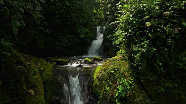 Drone flies low past multiple small cascading waterfalls dropping between moss-covered boulders into clear pools inside Mindo cloud forest, Ecuador