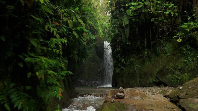 Drone glides above rocky rapids toward dramatic tall waterfall dropping into secluded dark pool enclosed by moss-covered canyon walls in Mindo forest, Ecuador