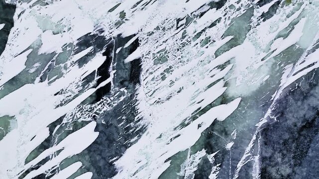 Aerial close-up of frozen sea surface with wind-swept snow streaks over dark blue slabs and turquoise patches, forming a dramatic natural abstract winter pattern.