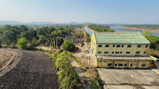 A static elevated view shows a hydroelectric powerhouse beside a switchyard, with a tailrace channel carrying discharged water through the landscape after power generation.