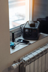 Naklejka premium Electricity backup at home in blackout, power outages, heating shutdown, energy crisis. Portable power station, power bank, small solar panel on window sill near home radiator. Vertical photo.