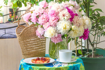 Beautiful large peony flowers bunch in transparent vase, cup of coffee or tea, strawberry cake on a table covered by tablecloth