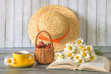 Summer still life; yellow cup of tea, bouquet of white daisy flowers on a open book, wicker basket with ripe strawberries and straw hat on a wooden background