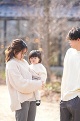 Fototapeta premium A woman is holding a baby while a man stands behind her. The scene is set in a park, with trees in the background. Scene is warm and friendly, as the family is spending time together outdoors