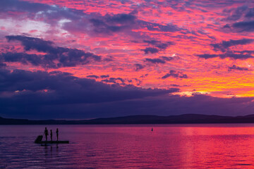 Sunset at Lake Taupō, New Zealand. 3 silhouetted swimmers on swim platform. Whangamatā Bay, Kinloch. Vivid pink, purple sky. © synthetick
