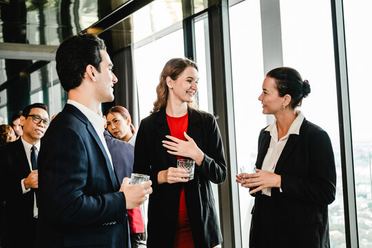 Professionals in business attire networking at a corporate event, chatting with drinks near large windows in a modern high-rise