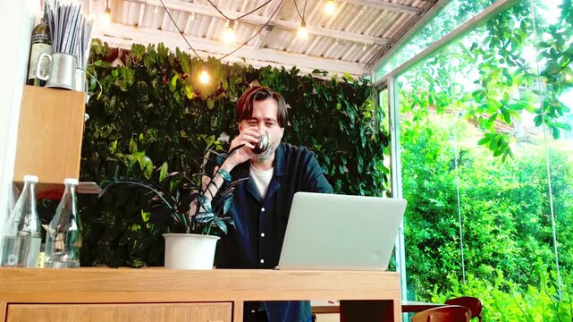 Person working on a laptop at a bright, plant-filled cafe patio, sipping coffee by a wooden counter with string lights and large windows, blending remote work with a relaxing green ambiance.