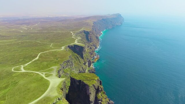 Aerial view of steep coastal cliffs and a shipwreck along the turquoise shoreline with green hills and dirt roads in Salalah, Oman.